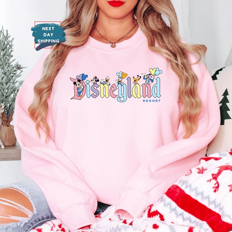以下が含まれることがあります： ディズニーリゾートのロゴの前にミッキーマウスとその仲間たちのグラフィックが描かれたピンク色のスウェットシャツです。
