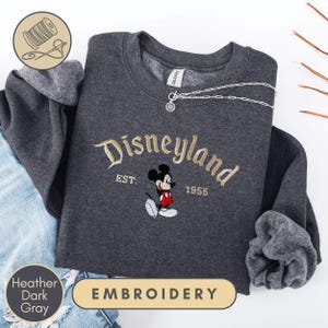 a gray sweatshirt with a Disney Mickey Mouse embroidered on it, along with a denim jacket and a pair of jeans. The sweatshirt has the text "Disneyland EST. 1955" and "Heather Dark Gray" printed on it.