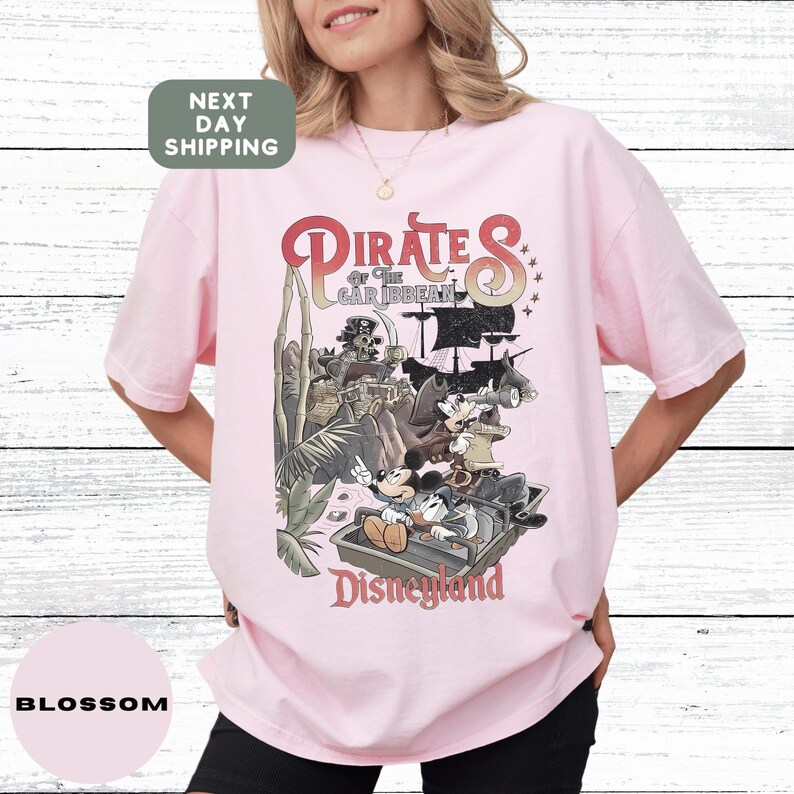 A woman wearing a pink t-shirt with a graphic of Disney characters and text related to the Pirates of the Caribbean ride at Disneyland.