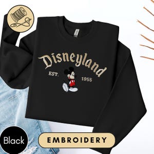 A black sweater with a Disneyland logo and text, along with a pair of blue jeans, is displayed on a white background.
