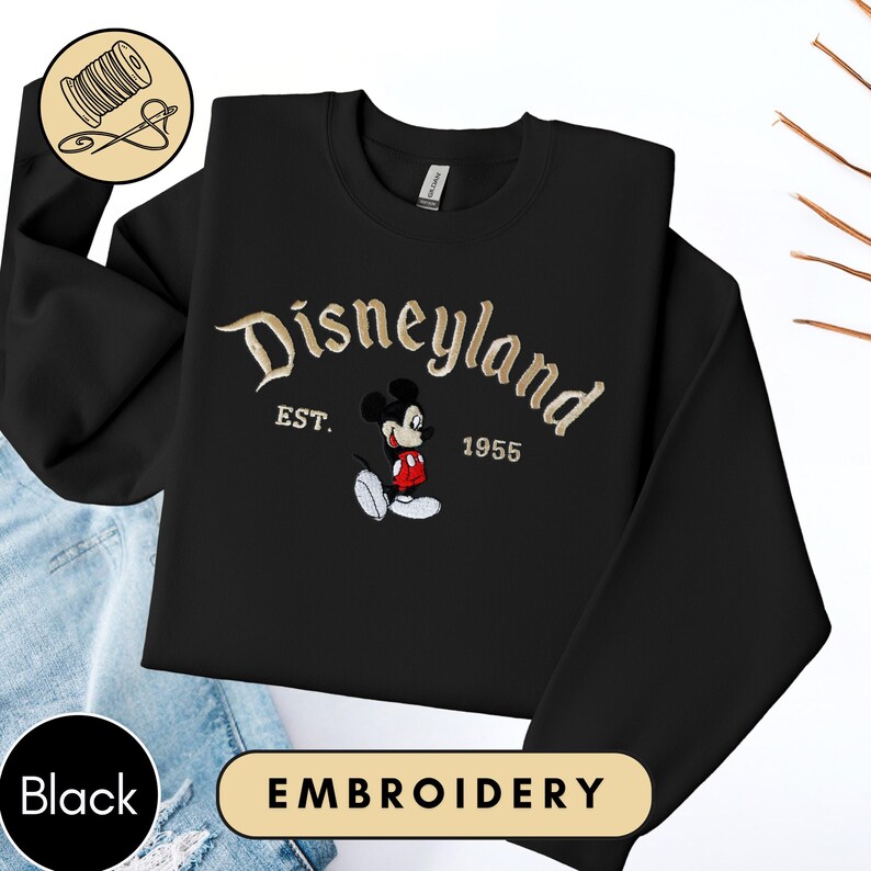 A black sweater with a Disneyland logo and text, along with a pair of blue jeans, is displayed on a white background.