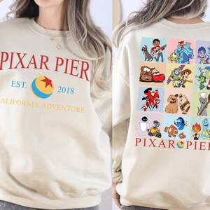May include: Cream-colored sweatshirt with "PIXAR PIER" in red lettering, a blue and yellow ball graphic, and the text "EST. 2018 CALIFORNIA ADVENTURE." The back features a grid of colorful Pixar character illustrations.