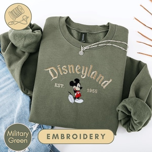A military green sweatshirt with a Disney Mickey Mouse embroidered on it, along with a chain and a spool of thread, indicating that it is an embroidery piece.