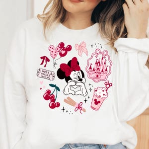 May include: White sweatshirt featuring a Minnie Mouse graphic with a red bow, surrounded by pink and red illustrations of balloons, a castle, cherries, and the text "Hugs & Kisses". The design has a Valentine's Day theme.
