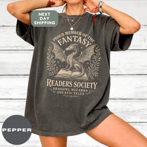 May include: Dark gray t-shirt with a fantasy-themed graphic. The design features a dragon on a book, surrounded by text that reads "Proud Member of the Fantasy Readers Society: Dragons, Wizards, and Epic Tales."