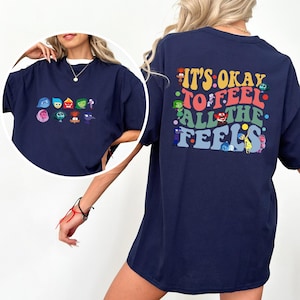 May include: Navy blue t-shirt featuring colorful cartoon characters and the text "IT'S OKAY TO FEEL ALL THE FEELS" on the back. The front of the shirt displays a collection of cartoon faces. A casual, oversized fit.