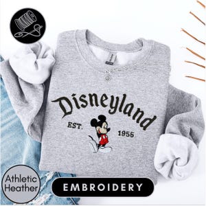 A gray sweatshirt with the text "Disneyland EST. 1955" and a Mickey Mouse embroidered on it, along with a spool of thread and a needle, suggesting the image is an embroidery of a Disney-themed sweatshirt.