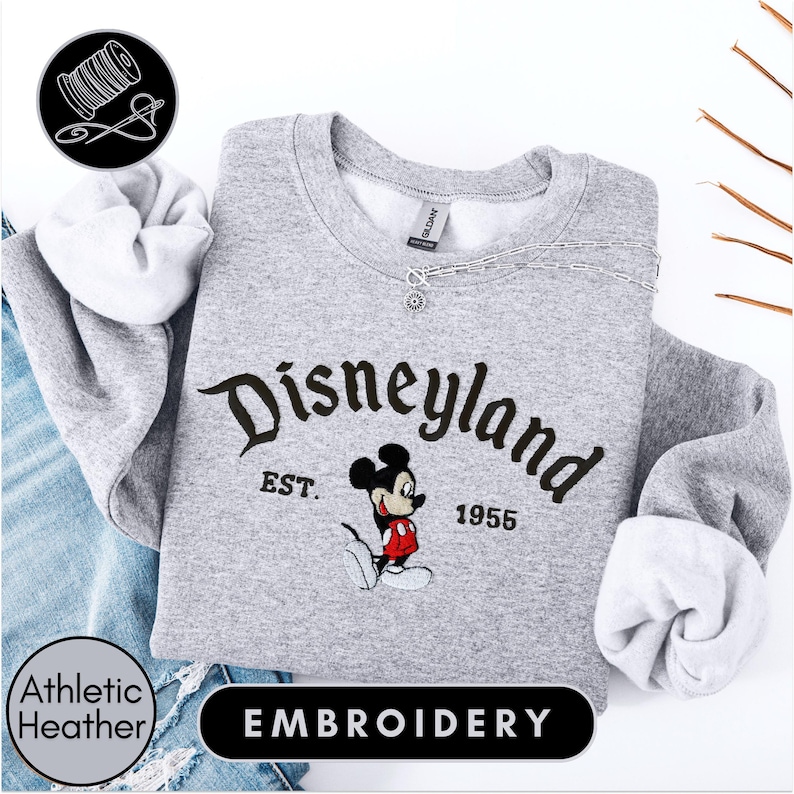 A gray sweatshirt with the text "Disneyland EST. 1955" and a Mickey Mouse embroidered on it, along with a spool of thread and a needle, suggesting the image is an embroidery of a Disney-themed sweatshirt.