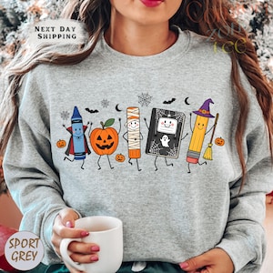 May include: A heather gray crewneck sweatshirt featuring a colorful graphic of five anthropomorphic school supplies dressed in Halloween costumes, including a crayon vampire, a jack-o'-lantern, a mummy glue stick, a ghost notebook, and a pencil witch.