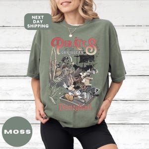 A woman wearing a green t-shirt with a graphic design of Disney's Pirates of the Caribbean on it, standing in front of a white wooden background.