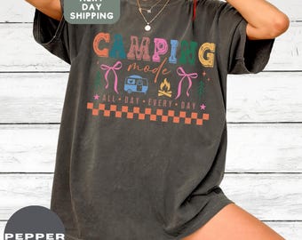 May include: Dark gray t-shirt with the words "CAMPING mode" in colorful letters. The shirt features a camper, campfire, and bow graphics. The text "ALL DAY EVERY DAY" is also included. The shirt is a relaxed fit.