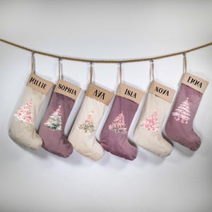 May include: Six Christmas stockings, each with a name: Millie, Sophia, Ava, Isla, Nova, and Emma. They hang from a rope and feature a Christmas tree design. The stockings are in beige and purple hues, perfect for festive decor.