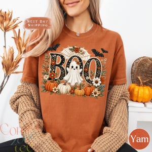 May include: A burnt orange t-shirt featuring a graphic print with the word BOO in large black letters decorated with floral patterns. A white ghost sits in the centre, surrounded by orange pumpkins, white flowers, spider webs, and black bats.