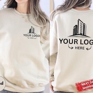 May include: A sand-colored crewneck sweatshirt featuring a black graphic of a building silhouette on the left chest and a larger version of the same graphic on the back. Both graphics include the black text "YOUR LOGO HERE" with arrows.