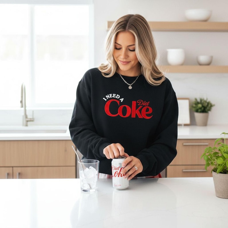 A woman in a black sweatshirt with the text "I NEED A Diet Coke" is standing at a kitchen counter, opening a can of Diet Coke.