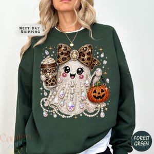 Peut inclure: Un sweat-shirt vert forêt orné d'un motif de fantôme décoré de fausses perles et cristaux. Le fantôme porte un nœud à motif léopard, tient une tasse à café léopard et transporte un seau citrouille orange avec une anse noire.