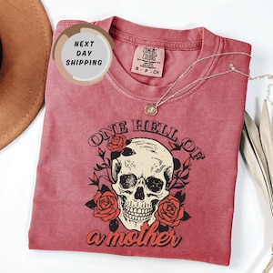 May include: A dusty rose Comfort Colors t-shirt featuring a skull graphic surrounded by roses and the text "ONE HELL OF A MOTHER". A silver necklace with a pendant rests on the shirt.