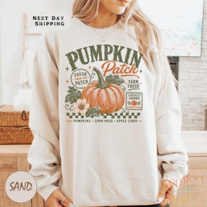 May include: A sand-colored crewneck sweatshirt featuring a graphic print of a large orange pumpkin. The text reads PUMPKIN Patch, FRESH FROM THE PATCH, FARM FRESH, LOCALLY GROWN, and PUMPKINS CORN MAZE APPLE CIDER. The design includes green leaves and stars.