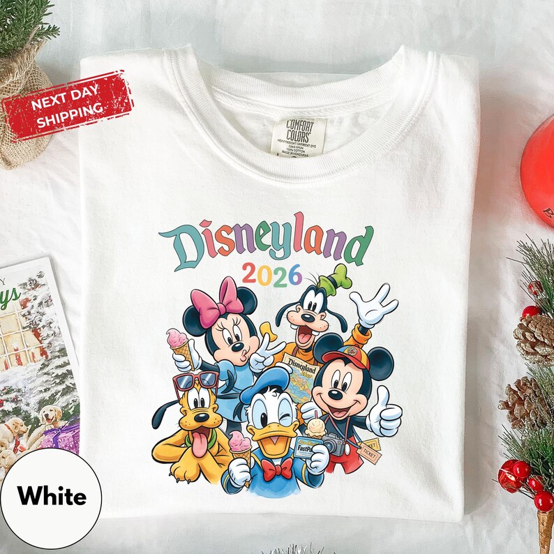 A white t-shirt with a colorful graphic of Disney characters from the theme park, including Minnie Mouse, Goofy, Donald Duck, and Mickey Mouse, along with the text "Disneyland 2026". The t-shirt is displayed on a white surface, surrounded by holiday decorations such as pine cones and ornaments.