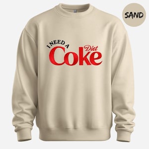 A beige sweatshirt with a red and black text design that says "I NEED A Diet Coke".