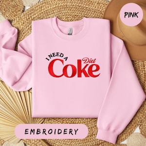 A pink sweatshirt with the text "I NEED A COCO Diet Coke" embroidered on it, along with a straw hat and a woven mat in the background.