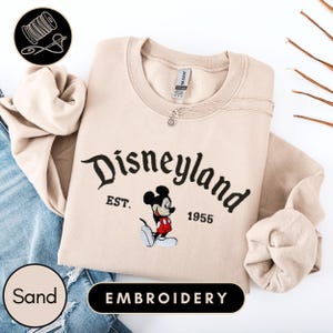 A beige sweater with the text "Disneyland EST. 1955" and an embroidered image of Mickey Mouse, along with the text "EMBROYDERY" below it.