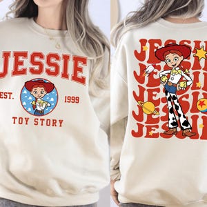 May include: A cream-colored sweatshirt featuring the name "JESSIE" in red block letters. The front design includes a circular graphic of the character Jessie from Toy Story, with "EST. 1999 TOY STORY" below. The back displays a larger Jessie graphic.