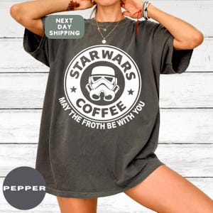 May include: Dark gray t-shirt with a white graphic featuring a Stormtrooper helmet inside a circle with the words "STAR WARS COFFEE" and "MAY THE FROTH BE WITH YOU". The shirt has a relaxed fit.