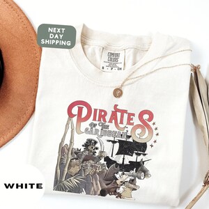 a white t-shirt with a pirate-themed graphic design, including a pirate ship, pirate characters, and other pirate-related imagery.