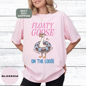 May include: A light pink t-shirt with the words "FLOATY GOOSE" in pink and blue text. The shirt features a cartoon goose wearing sunglasses and a swim ring. The text "ON THE LOOSE" is below the goose.