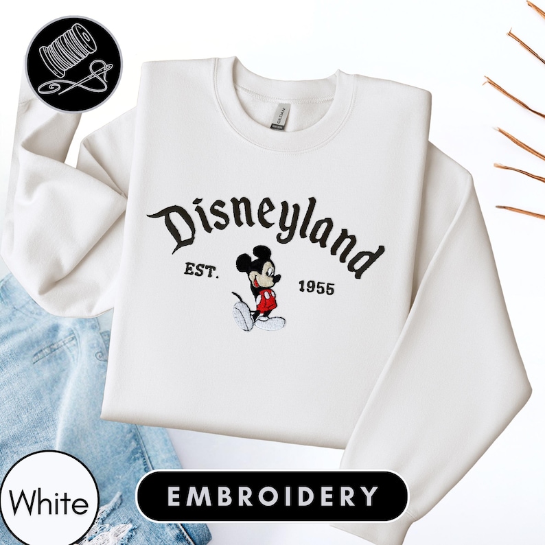 A white sweatshirt with the text "Disneyland EST. 1955" and an embroidered image of Mickey Mouse, a classic Disney character.