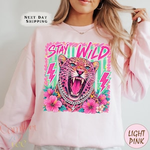 May include: A light pink sweatshirt featuring a graphic of a leopard with an open mouth, wearing a silver chain necklace. The design includes the text STAY WILD, pink lightning bolts, and pink hibiscus flowers against a green striped background.