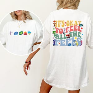 May include: White t-shirt featuring colorful cartoon characters and the text "IT'S OKAY TO FEEL ALL THE FEELS" on the back. The front of the shirt displays the cartoon characters. The shirt is a casual, oversized fit.