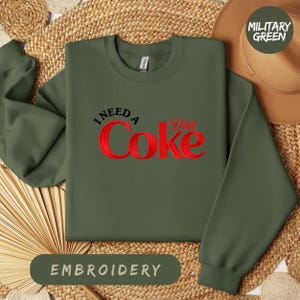 a green sweatshirt with a red Coca Cola logo, laid out on a woven mat. The sweatshirt has a military green color, and there is a hat placed next to it. The image also includes the text "I NEED A Diet Coke" and "EMBROYERY" in the corners.