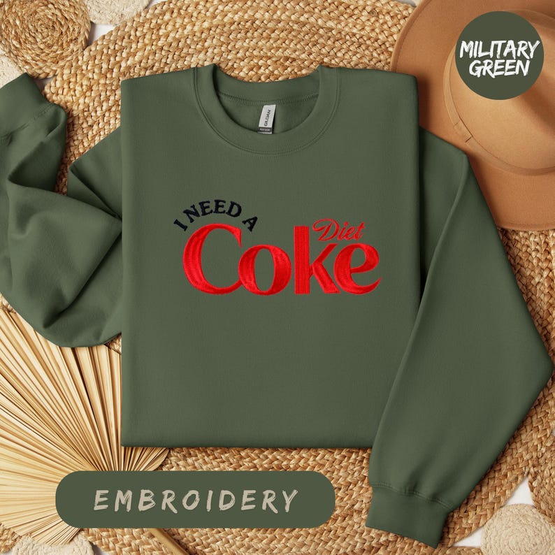 a green sweatshirt with a red Coca Cola logo, laid out on a woven mat. The sweatshirt has a military green color, and there is a hat placed next to it. The image also includes the text "I NEED A Diet Coke" and "EMBROYERY" in the corners.