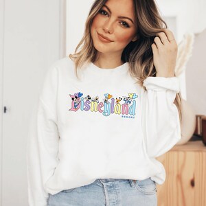 以下が含まれることがあります： ミッキーマウスとその仲間たちが描かれたカラフルなディズニーランドリゾートのロゴがプリントされた白い長袖スウェットシャツ。ロゴはパステルカラーと風船を使ったヴィンテージスタイルです。