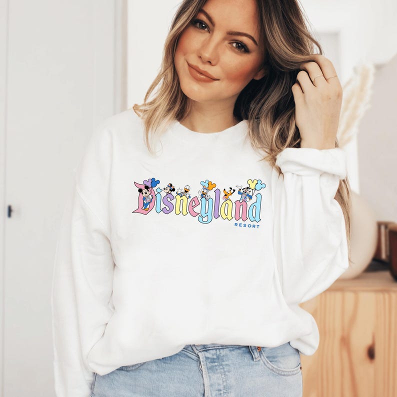 以下が含まれることがあります： ミッキーマウスとその仲間たちが描かれたカラフルなディズニーランドリゾートのロゴがプリントされた白い長袖スウェットシャツ。ロゴはパステルカラーと風船を使ったヴィンテージスタイルです。