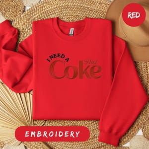 a red sweatshirt with the text "I NEED A Diet Coke" printed on it, along with a straw hat and a woven mat in the background.