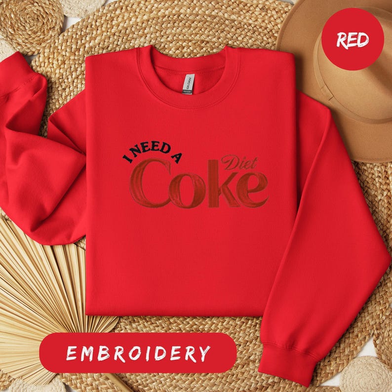 a red sweatshirt with the text "I NEED A Diet Coke" printed on it, along with a straw hat and a woven mat in the background.