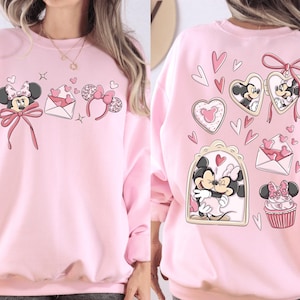 May include: Pink sweatshirt featuring Minnie Mouse and Valentine's Day graphics. The front displays Minnie Mouse, an envelope with hearts, and a headband. The back showcases Minnie and Mickey Mouse in heart-shaped frames, hearts, and a cupcake.