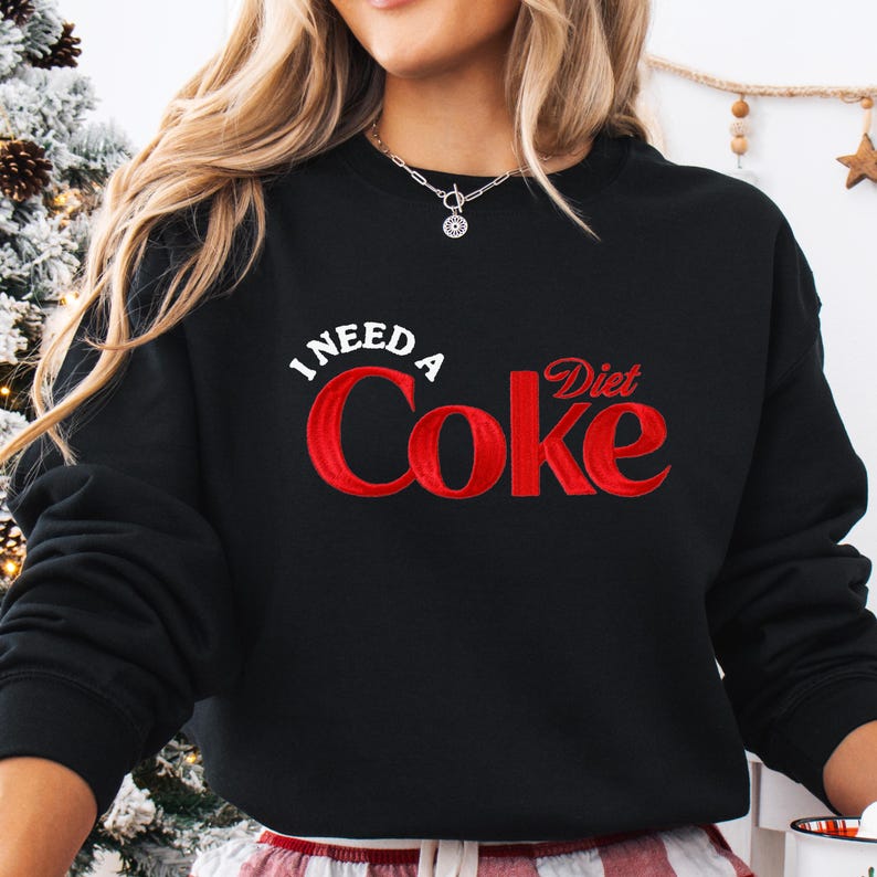 A woman wearing a black sweatshirt with the text "I NEED A Diet Coke" printed on it.