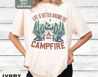 May include: Ivory-colored t-shirt with a camping-themed graphic. The design features the text "LIFE IS BETTER AROUND THE CAMPFIRE" above a campfire scene with two chairs, trees, mountains, and a crescent moon. The shirt has short sleeves.