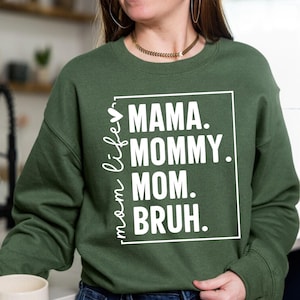 May include: Dark green sweatshirt with the words "MAMA. MOMMY. MOM. BRUH." in a white rectangular box. The words "mom life" are written vertically in white on the left side of the sweatshirt. The sweatshirt is a crew neck style.