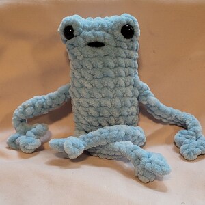 May include: A light blue crocheted frog plush toy with black eyes. The frog is sitting with its legs outstretched.