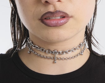 May include: A silver chain choker necklace with two chains and a cross pendant. The necklace has small ball accents on the ends of the chains.