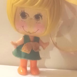Pode incluir: Uma boneca pequena com cabeça grande, cabelo moldado amarelo e traços faciais pintados, incluindo bochechas rosadas. A boneca veste um vestido verde com um laço bege na cintura e botas laranja brilhantes. Uma fita amarela está presa na parte de trás da cabeça.