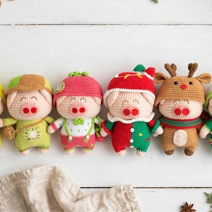 Op de afbeelding: Zes gehaakte varkenspoppen op een rij, elk met een andere outfit. De varkens hebben lichamen van lichtroze garen, ronde snuiten en gesloten ogen. De outfits omvatten een ananas-hoed, een groene pet, een aardbeien-hoed, een rood-groen pak, een rendier-capuchon en een witte hoed met feestelijke versiering.