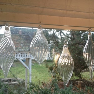 Könnte beinhalten: Fünf klare Glaskugeln hängen an einem Fenster. Die Kugeln sind tropfenförmig und haben ein Spiralmuster. Sie hängen vor einem grünen, belaubten Hintergrund.