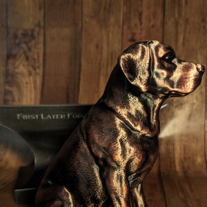 May include: A 3D printed bronze-coloured dog figurine sitting on a wooden surface. The dog is in a sitting position with its head turned to the side. The figurine is detailed and realistic.