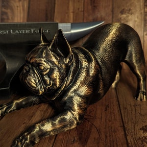Puede incluir: Una figurita de un Bulldog Francés acostado, impresa en 3D en color dorado. La figurita es detallada y tiene una textura realista.